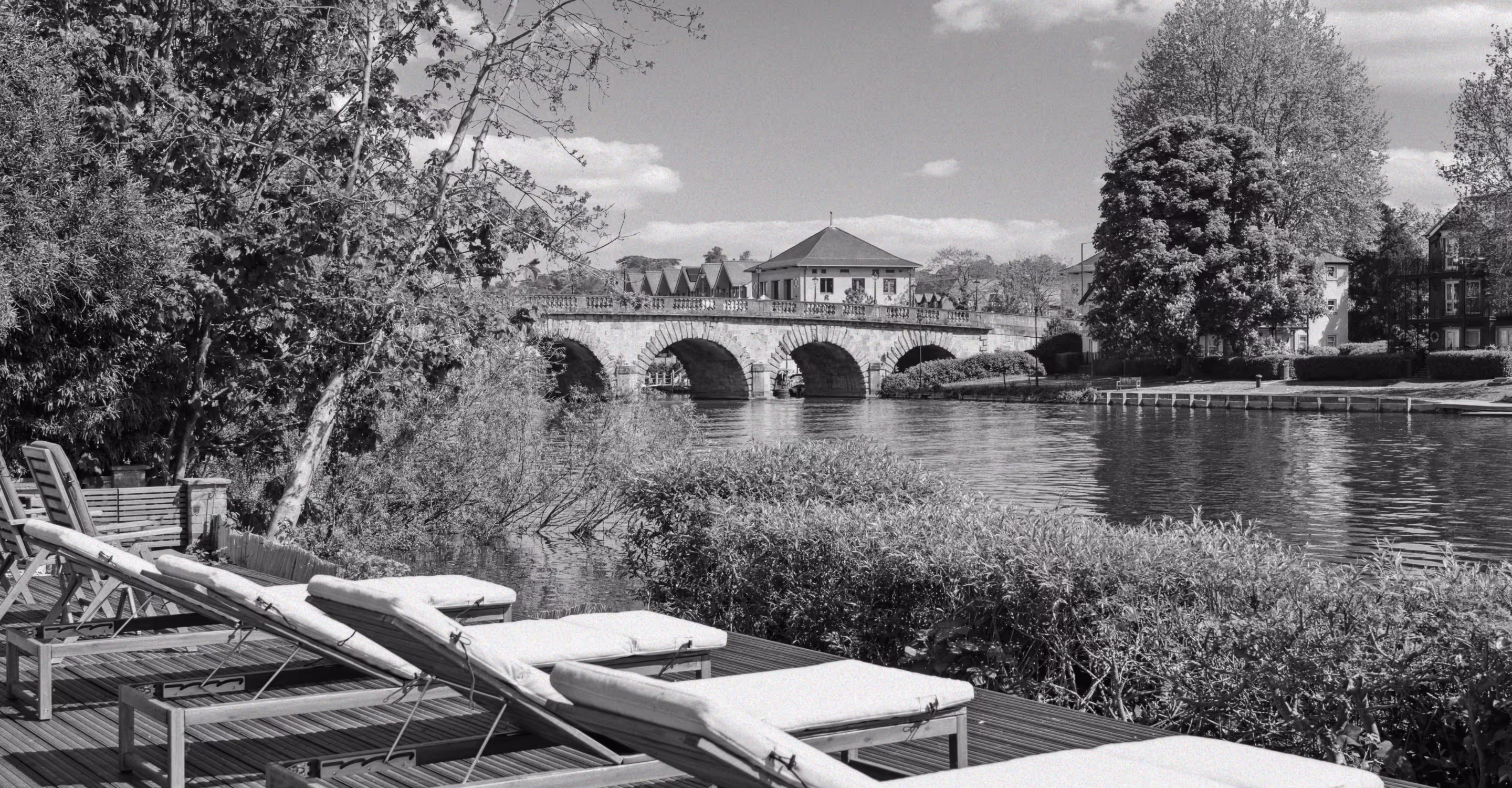 Black-and-white view of sun loungers on a riverside deck looking across the Thames to a stone bridge.
