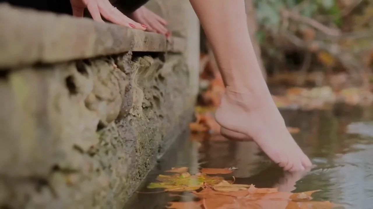 Bare feet dipping into the river from a mossy stone edge, autumn leaves on the water