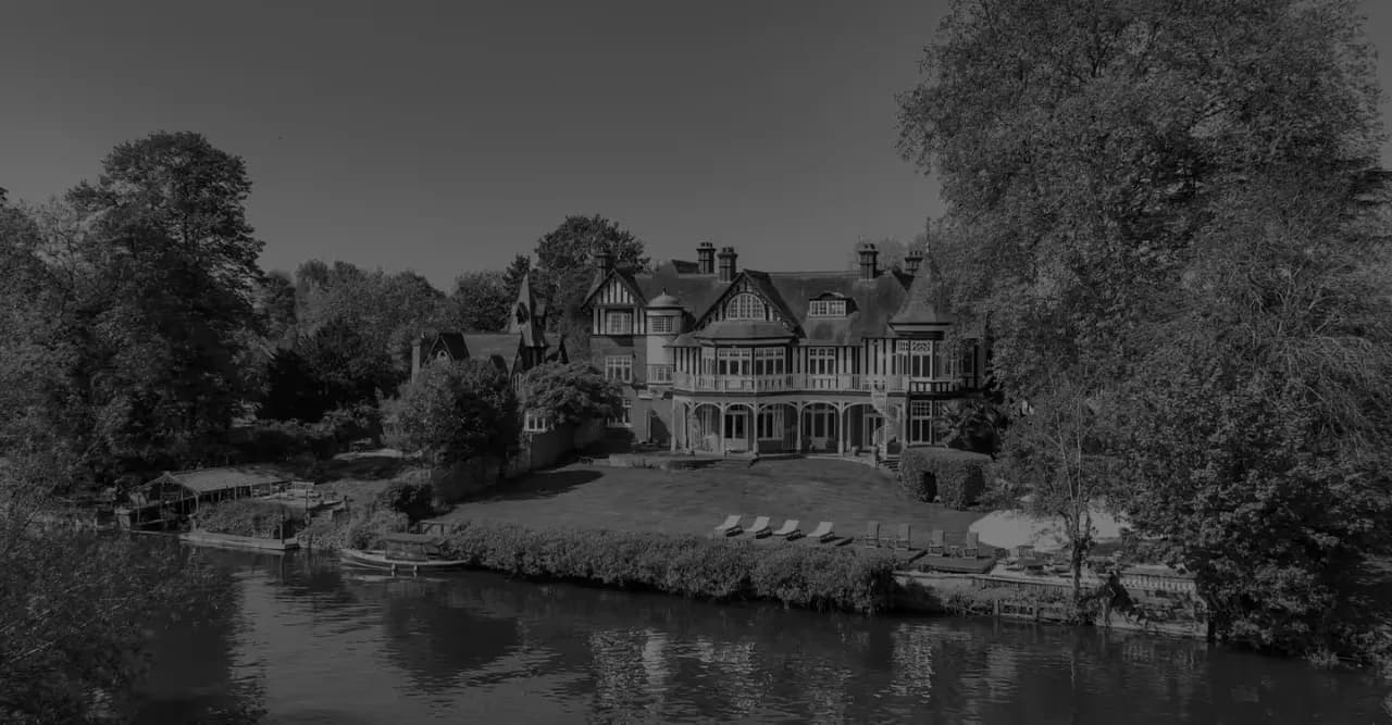 Black-and-white view of a Tudor-style riverside manor house with lawn and boat dock, seen across the river.